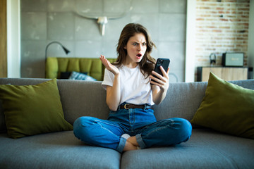Portrait of a surprised woman holding a phone looking at you sitting on a sofa in the living room