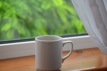 A white cup of tea or coffee stands on a wooden window sill against the background of a summer garden