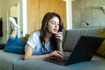 Beautiful young woman working from home on a laptop lying on the sofa in the room