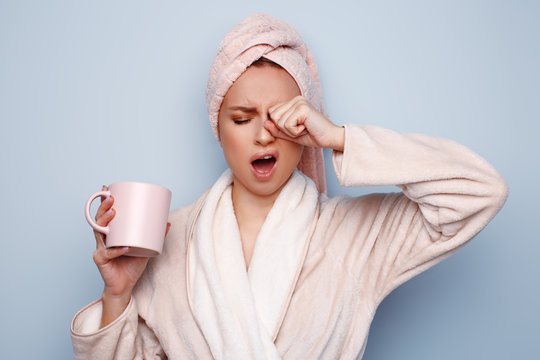 Woman In Pink Bathrobe With Towel On Head Holding Cup Of Tea Or Coffee In Hand, After Shower At Morning, Sleepy And Tired, Wants To Sleep, Rubbing Her Eyes And Yawning. Shift Work Sleep Disorder