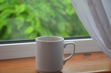 A white cup of tea or coffee stands on a wooden window sill against the background of a summer garden