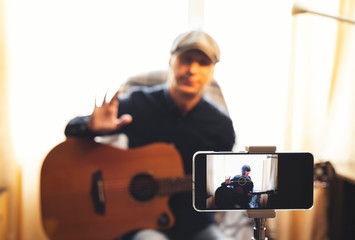 Man with acoustic guitar in a shirt and cap plays live staying home for the audience in front of the smartphone camera during quarantine and forced self-isolation due to coronavirus