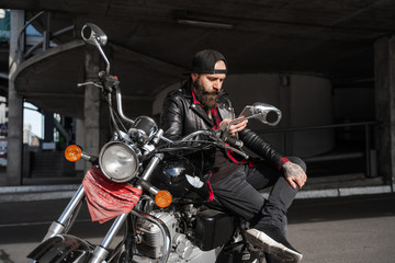 Fototapeta na wymiar Brutal bearded biker man sitting on bike in black with phone, motorcycle style, stylish man using mobile cell phone and Internet technologies online, communication via 5g connection. 