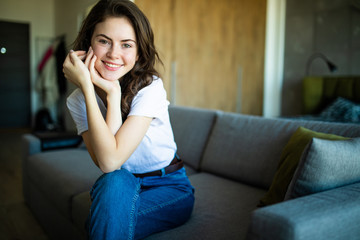 Obraz premium Young dreaming woman sitting on sofa at home. Thoughtful young woman sitting in living room and looking up. Smiling woman relaxing on couch in a modern home.