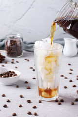 Cold coffee is poured into a tall glass with ice cubes on light gray background, Closeup