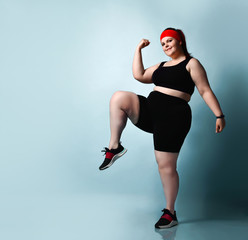 Fototapeta na wymiar Active smiling plus-size girl practices exercises stepping in place raising up knees high on blue background. Fitness Full length portrait, copy space