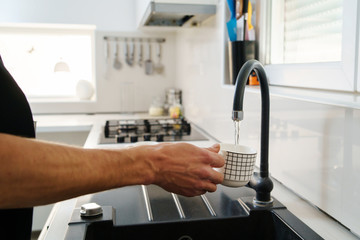 Close up on male hands unknown caucasian man holding white ceramic cup at sink tap faucet pouring water in the kitchen at home in day side view close up