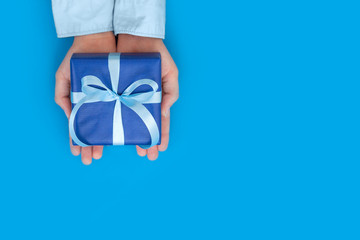 Kid's hands holding gift box wrapped in craft paper and tied with bow on blue background. Concept Birthday or Father's Day.