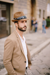 A young guy with a straw hat, a white unbuttoned shirt and a sand jacket, a small beard walks around the city of Florence.