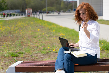Fototapeta premium Red-haired woman in a park on a bench works in a laptop and makes notes with a pencil in a diary
