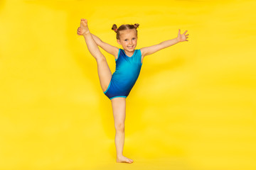 Young cute barefooted gymnast girl with two buns in blue sport suit posing on yellow background...