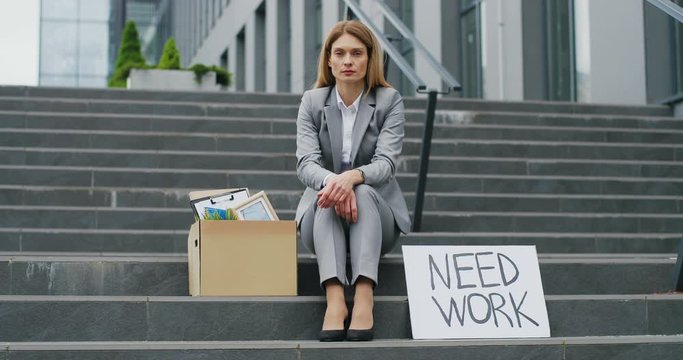 Portrait of young workless Caucasian woman office worker sitting on stairs outdoors with carton table Need Work. Unemployment after pandemic lockdown. Workless fired female.