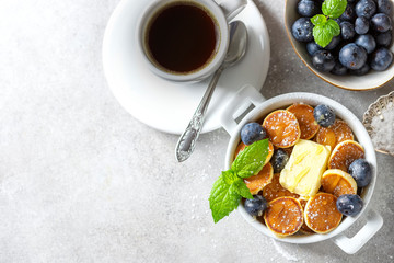 Bowl with tiny pancake cereal on a light concrete background. Trendy food. Mini cereal pancakes with butter, honey and blueberries.