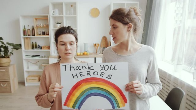 Two young women showing rainbow painting with inscription «NHS thank you heroes», looking at camera and saying thanks to healthcare workers while staying at home. Covid-19 pandemic concept