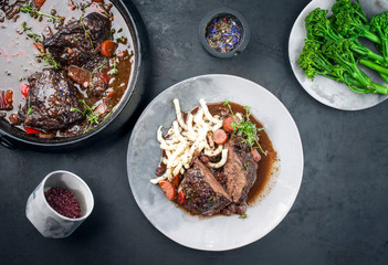 Traditional German braised beef cheeks in brown red wine sauce with noodles and carrots offered as top view on a modern design plate and stewpot on an old rustic board