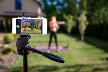 Sportive fitness coach woman recording video blog by phone about workout training at the garden with fresh green grass near house.