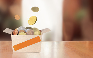 A carton donation box with an orange adhesive tape strip on a table full of money with a blurred window behind. Falling coins levitation. Local business support concept.
