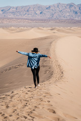 A travel girl is jumping on the top of a sand dune back to the camera. A traveler in a black hat...