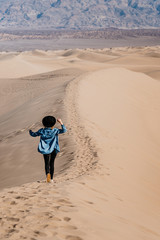 A travel girl is going on the top of a sand dune back to the camera. A traveler in a black hat and blue skirt in Death Valley National Park