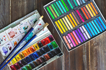 Flat lay art workplace, wooden table with paint brushes, watercolor paints and crayon pastel chalks close up. Copy space