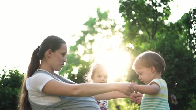 family, children, motherhood, entertainment and people concept - young cute mother of many children with her baby in sling plays with two older kids same age on background of the sunset in the park.