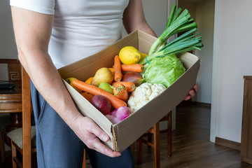 Obraz premium A volunteer, a food delivery man is holding a box of vegetables. Fresh vegetables in cardboard box on brown wood background.