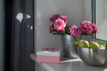 bouquet of red roses, a steel bowl with green apples and a pink box with the inscription Best wishes on a stone windowsill. Room interior with gray curtains