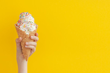 Children's hand holds delicious vanilla ice cream cone on a bright yellow background.