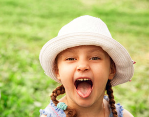 Fun kid girl in fashion hat looking excited her big eyes with humor face with opened mouth  on summer green grass background. Closeup