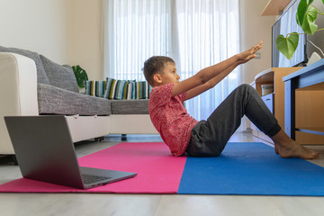 Teenager boy with laptop computer doing sport exercises, practicing yoga in the living room. Sport, healhty lifestyle, active leisure at home