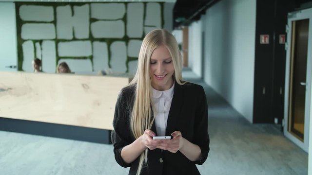 Stylish blond woman in jacket standing in contemporary office hall and surfing smartphone with smile