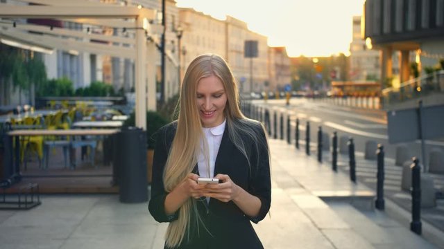 Elegant blond woman in suit walking on street and browsing smartphone with smile against urban background.