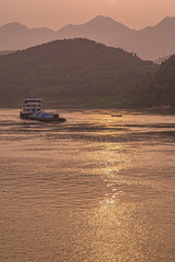 Chongqing, China - May 8, 2010: Evening light on Yangtze River. Loaded barge sails on while golden sunset reflected by brown water in front of shoreline with forested hills. Small fishing sloop presen