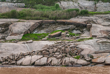 Chongqing, China - May 8, 2010: Evening light on Yangtze River. Closeup of geology of rocky shoreline with patches of green grass and weeds behind brown water.