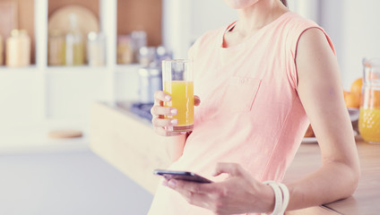 Obraz na płótnie Canvas Smiling pretty woman looking at mobile phone and holding glass of orange juice while having breakfast in a kitchen.