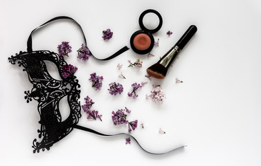 A composition of a masquerade black mask, spring flowers and tassels on a white background. Purple flowers are scattered randomly