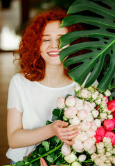 Portrait of a smiling red-haired curly girl with a bouquet of spring flowers in her hands, hiding part of her face behind a green leaf with a flowerpot