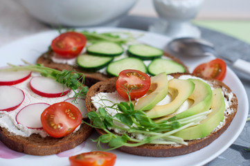 Vegetarian sandwiches with cream cheese, cucumber, avocado, garden radish, tomatoes and peas sprouts on white plate. Egg mist on white stand. Breakfast with vegetarian food.