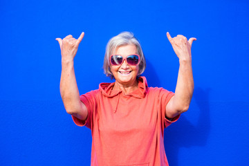 close up and portrait of funny and crazy senior or mature woman doing surfing sign and smililing looking at the camera with blue background - pensioner wearing red clothes and sunglasses happy people