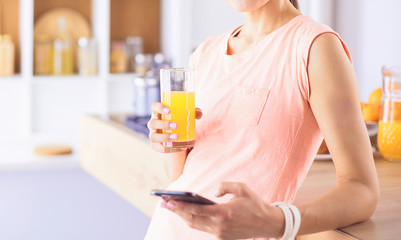 Smiling pretty woman looking at mobile phone and holding glass of orange juice while having breakfast in a kitchen.