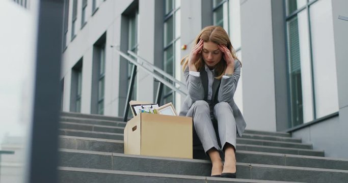 Fired Caucasian femae office worker sitting on stairs in depression with box of stuff. Unemployed businesswoman lost her business. Anxious concept. Workless woman in despair.