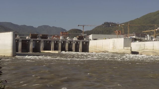 Panning shot of river flowing from dam with cranes near mountain against sky on sunny day - Luang Prabang, Laos