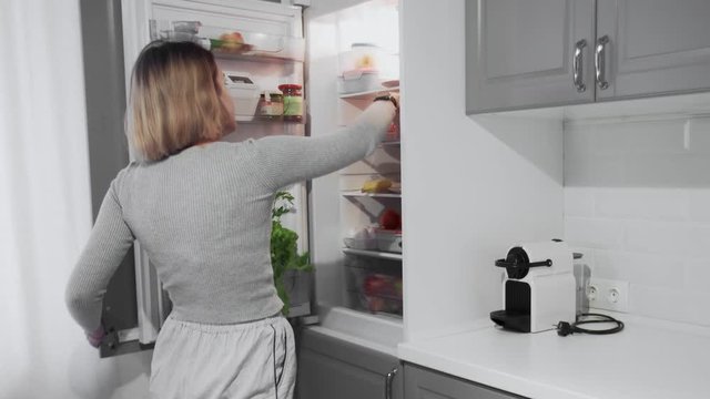 Woman opens refrigerator door in kitchen at home, takes apple then puts it back and takes donut