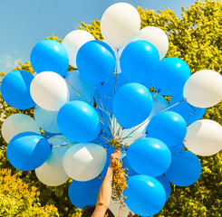 Male hand holding bunch of blue and white balloons against green tree and blue sky on sunny day. Holiday, celebration, Children's Day, wedding, graduation concept. Peace, love, freedom, purity idea.