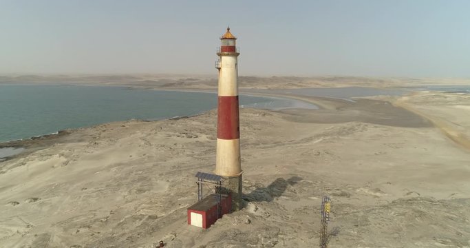Aerial shot of tall lighthouse at beach near sea against sky, drone panning from left to right over rock formation on sunny day - Luderitz, Namibia