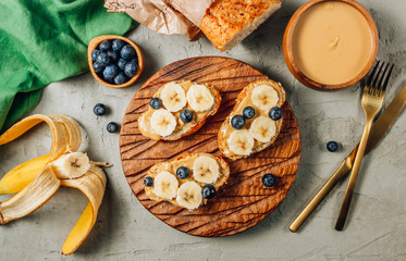 Buckwheat healthy bread with peanut butter, banana and blueberry on wooden board over concrete background. Top view. Flat lay. Summer breakfast.