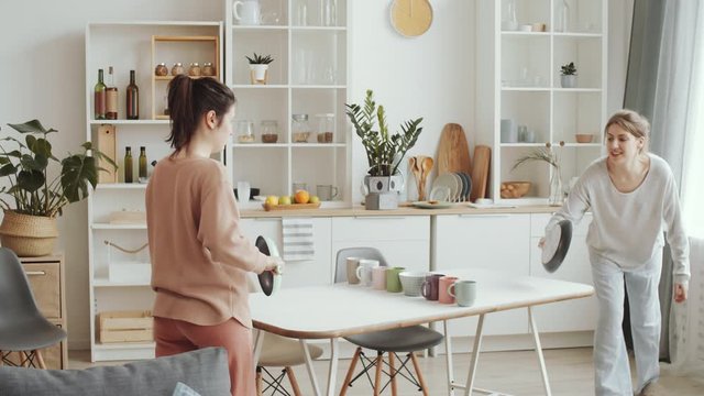 Two Young Female Friends Playing Ping Pong With Frying Pans As Racquets And Tea Cups Instead Of Net At Kitchen Table While Spending Time Together At Home Under Lockdown