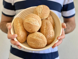 A person holds in his hands a basket with small round and oval home-made breakfast buns from whole...