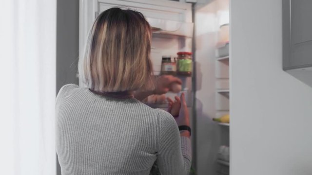 Woman opens refrigerator door in kitchen at home and takes two eggs and bottle of milk