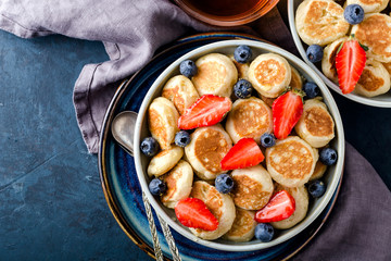 Trendy food - pancake cereal. Mini cereal pancakes with strawberries and blueberries in boul on blue cement background, top view. Copy space left for text.